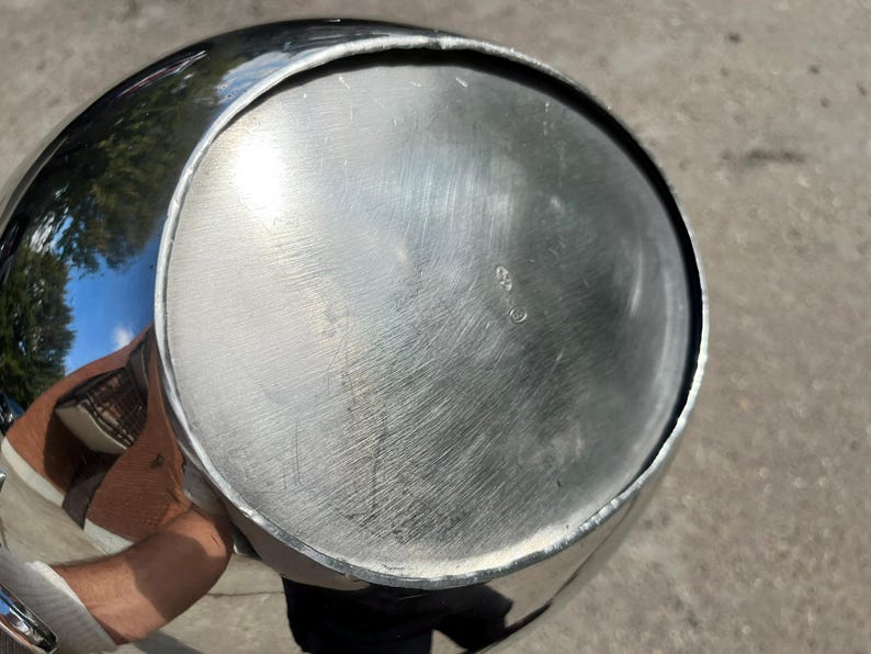 Puede incluir: Un recipiente de metal plateado brillante con forma redondeada. La parte inferior del recipiente tiene un dise&ntilde;o circular con ara&ntilde;azos visibles. El recipiente refleja el entorno, incluidos &aacute;rboles y cielo.