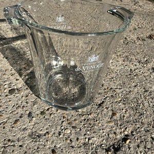 May include: A clear glass champagne bucket with two handles. The bucket has the words "de Venoge" printed on the front. The bucket is on a grey concrete surface.