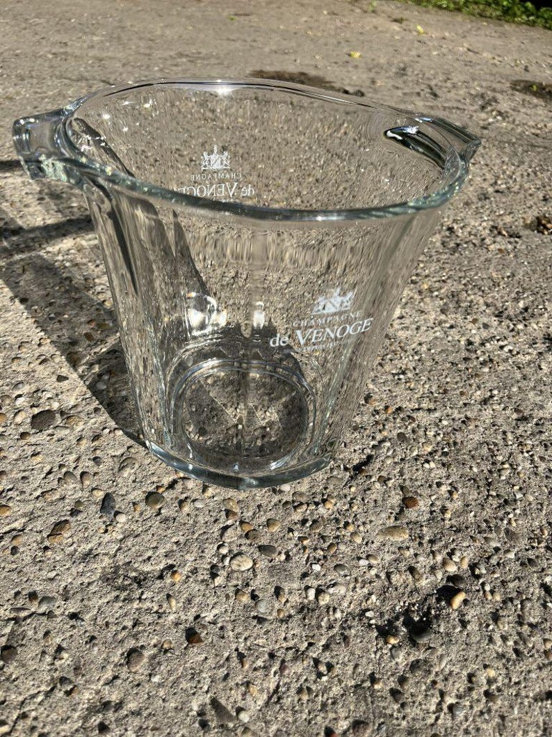 May include: A clear glass champagne bucket with two handles. The bucket has the words "de Venoge" printed on the front. The bucket is on a grey concrete surface.