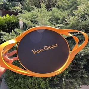 Puede incluir: Una bandeja de servicio Veuve Clicquot negra y naranja. La bandeja tiene un centro circular negro con el nombre de la marca en letras doradas. La bandeja tiene un borde y un asa de color naranja. La bandeja se sostiene frente a un arbusto verde.