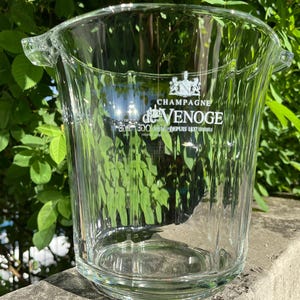 May include: A clear glass champagne bucket with the text "Champagne de Venoge" in white. The bucket has a ribbed design and two handles. The background features green foliage.