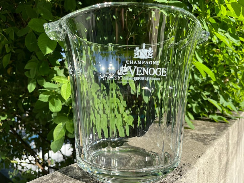 May include: A clear glass champagne bucket with the text "Champagne de Venoge" in white. The bucket has a ribbed design and two handles. The background features green foliage.
