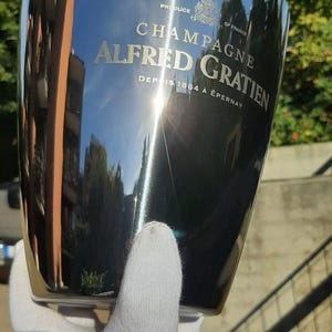 Pode incluir: Um balde de champanhe em a&ccedil;o inoxid&aacute;vel polido com as palavras "CHAMPAGNE ALFRED GRATIEN" e "PRODUCE OF FRANCE". O balde &eacute; segurado por uma m&atilde;o enluvada e reflete o ambiente. O texto "DEPUIS 1864 &Agrave; &Eacute;PERNAY" tamb&eacute;m &eacute; vis&iacute;vel.