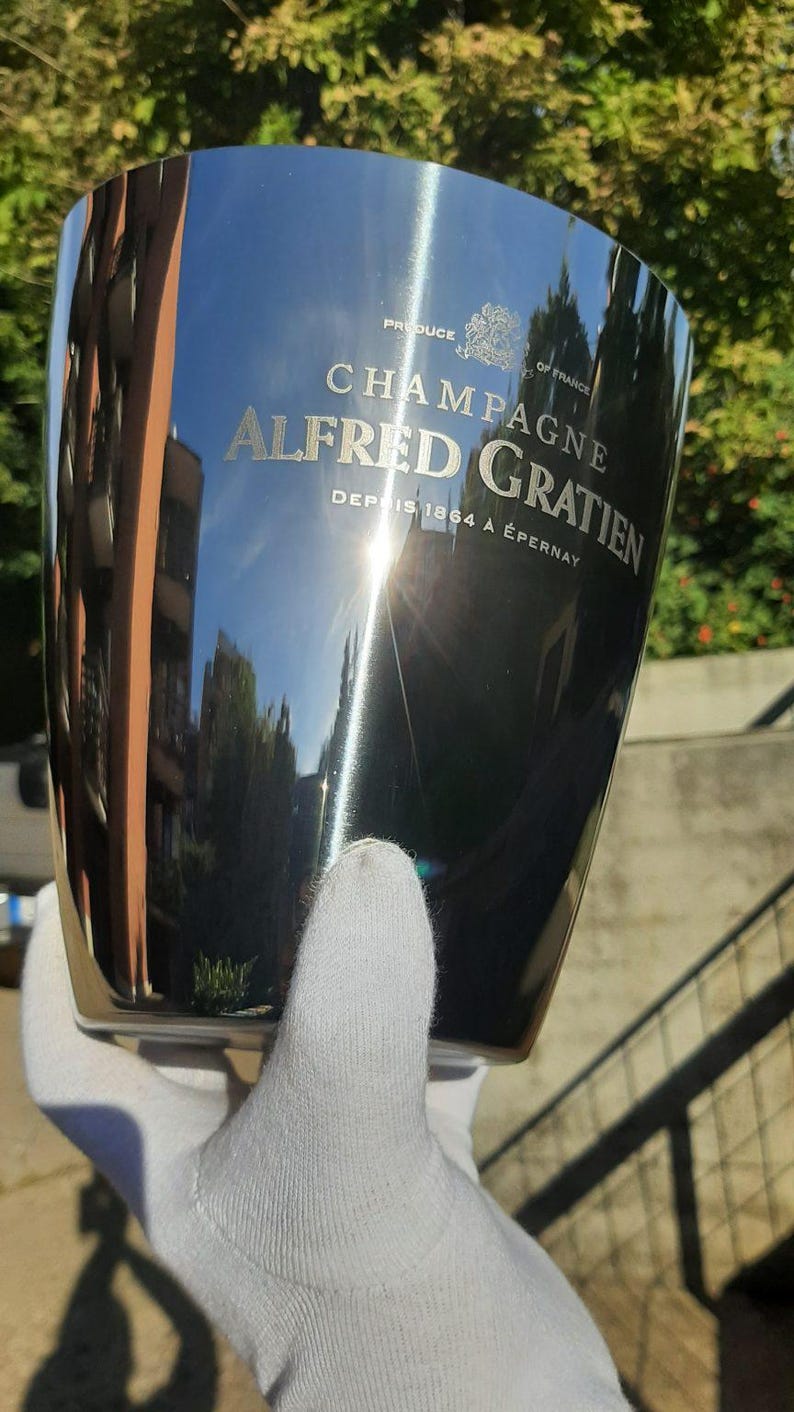Pode incluir: Um balde de champanhe em a&ccedil;o inoxid&aacute;vel polido com as palavras "CHAMPAGNE ALFRED GRATIEN" e "PRODUCE OF FRANCE". O balde &eacute; segurado por uma m&atilde;o enluvada e reflete o ambiente. O texto "DEPUIS 1864 &Agrave; &Eacute;PERNAY" tamb&eacute;m &eacute; vis&iacute;vel.