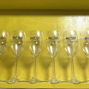 May include: Six clear champagne flutes with the "Ruinart Champagne" logo printed in black. The flutes are arranged in a row inside a yellow box. The glasses have a tall, slender design.