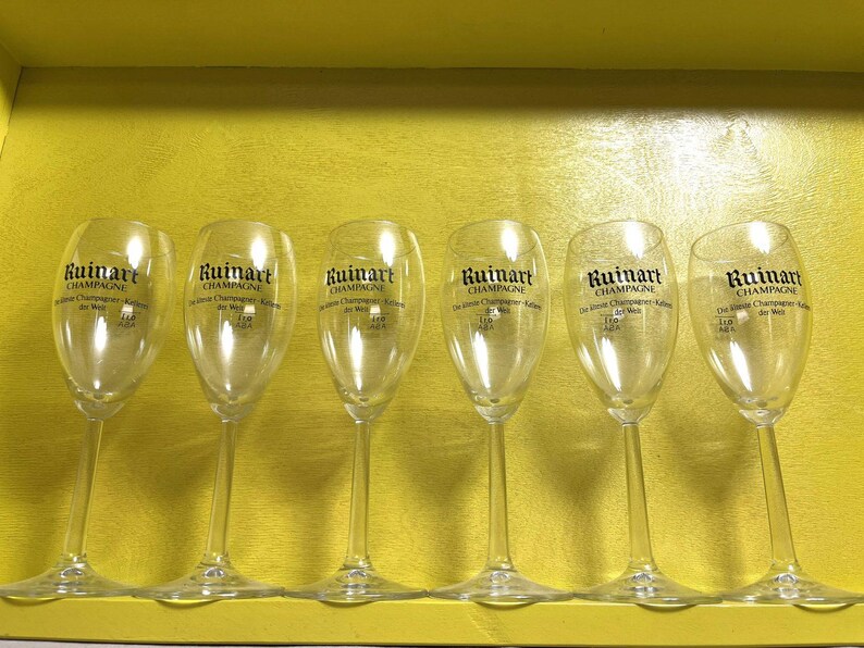 May include: Six clear champagne flutes with the "Ruinart Champagne" logo printed in black. The flutes are arranged in a row inside a yellow box. The glasses have a tall, slender design.