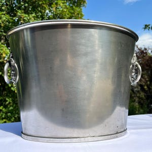 Puede incluir: Una cubitera grande de metal cepillado con asas decorativas. La cubitera tiene una forma cilíndrica ancha y un borde ligeramente ensanchado. Las asas son ornamentadas, con un diseño detallado. La cubitera está sobre una superficie blanca, con un fondo verde y azul.