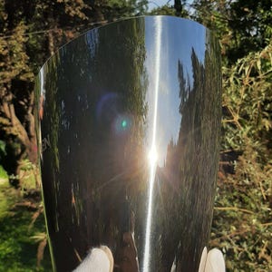 Pode incluir: Um balde de vinho de a&ccedil;o inoxid&aacute;vel brilhante com um design curvo. A superf&iacute;cie reflete as &aacute;rvores e o c&eacute;u circundantes, com um brilho solar brilhante. O balde &eacute; segurado por uma m&atilde;o enluvada, sugerindo um foco na apresenta&ccedil;&atilde;o e eleg&acirc;ncia.