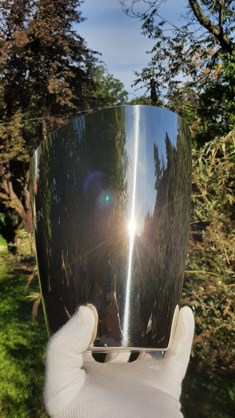 Pode incluir: Um balde de vinho de a&ccedil;o inoxid&aacute;vel brilhante com um design curvo. A superf&iacute;cie reflete as &aacute;rvores e o c&eacute;u circundantes, com um brilho solar brilhante. O balde &eacute; segurado por uma m&atilde;o enluvada, sugerindo um foco na apresenta&ccedil;&atilde;o e eleg&acirc;ncia.