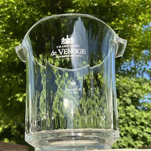 May include: A clear glass champagne bucket with the text "CHAMPAGNE de VENOGE" in white. The bucket has a curved top with two handles and a ridged design. The background is a blurred view of green foliage.