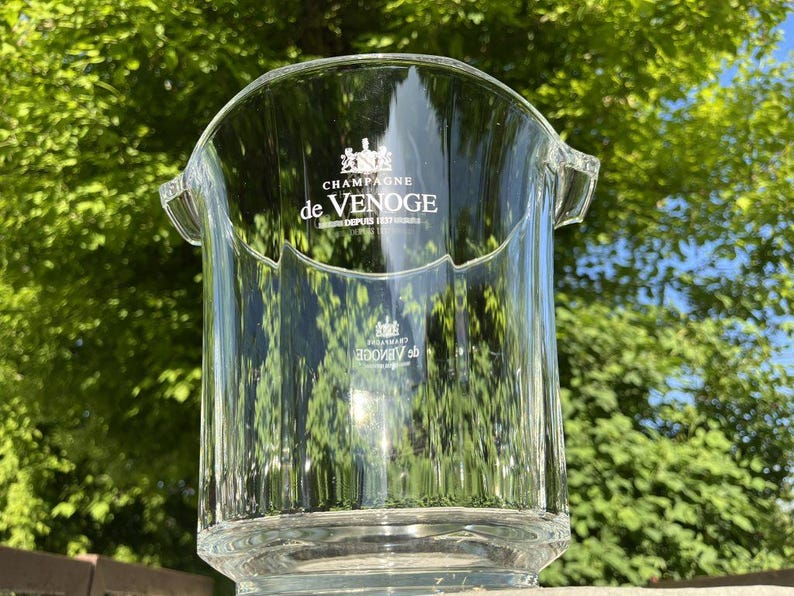 May include: A clear glass champagne bucket with the text "CHAMPAGNE de VENOGE" in white. The bucket has a curved top with two handles and a ridged design. The background is a blurred view of green foliage.