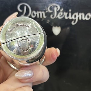 Könnte beinhalten: Ein silberfarbener, kugelförmiger Flaschenverschluss mit dem Dom Pérignon-Logo auf der Oberseite. Der Verschluss hat ein Scharnierdesign und ein Schildemblem. Der Hintergrund zeigt das Dom Pérignon-Logo in Weiß.