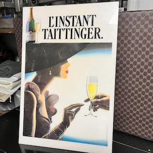 May include: A vintage advertisement for Taittinger champagne. The artwork features a woman in a large hat and gloves, holding a champagne glass. The text "L'INSTANT TAITTINGER" is displayed at the top, with a bottle and glass of champagne on the left.