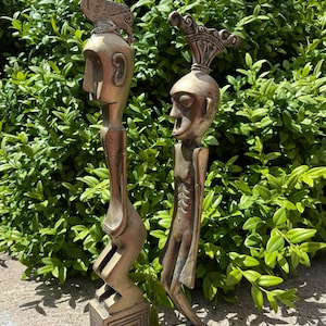 May include: Two bronze-coloured sculptures of stylised human figures. One figure has a bird on its head, the other has a crown-like structure. Both figures stand on decorative bases, set against a backdrop of green foliage.