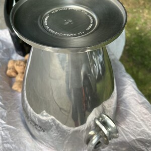 Puede incluir: Un enfriador de vino de metal plateado con una parte superior ancha y plana y un cuerpo bulboso. El enfriador tiene un asa y est&aacute; sobre una superficie blanca. Una botella oscura y corchos de champ&aacute;n est&aacute;n en el fondo.