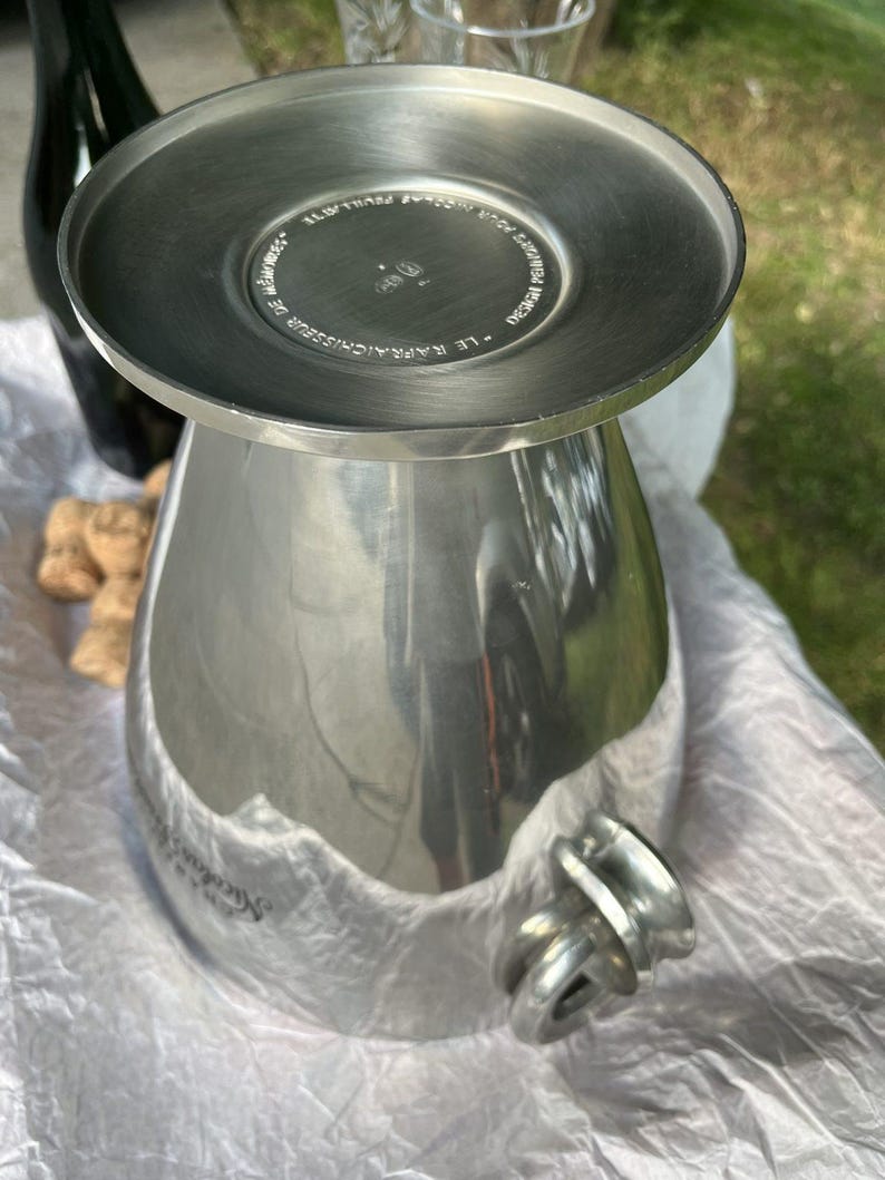 Puede incluir: Un enfriador de vino de metal plateado con una parte superior ancha y plana y un cuerpo bulboso. El enfriador tiene un asa y est&aacute; sobre una superficie blanca. Una botella oscura y corchos de champ&aacute;n est&aacute;n en el fondo.