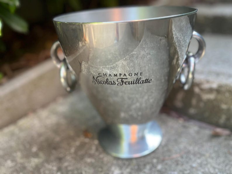 Puede incluir: Una cubeta de champ&aacute;n plateada con dos asas de anillo. La cubeta tiene las palabras "CHAMPAGNE Nicolas Feuillatte" impresas en la parte delantera. La cubeta est&aacute; sobre una superficie de piedra.