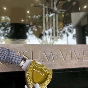 K&ouml;nnte beinhalten: Ein schildf&ouml;rmiger, olivgr&uuml;ner Keramik-Aschenbecher mit dem Logo "Mo&euml;t & Chandon". Der Aschenbecher wird von behandschuhten H&auml;nden vor einer Steinmauer mit der Inschrift "SUM VIVE" gehalten. Der Aschenbecher hat ein dekoratives Weinranken-Design.