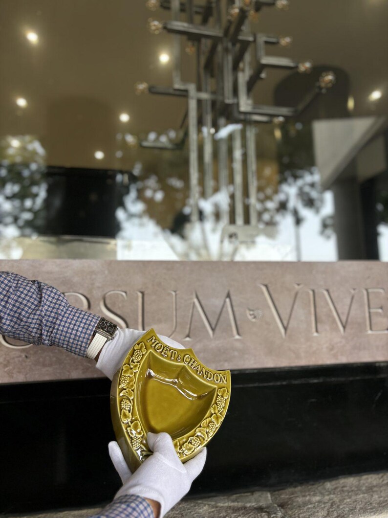 K&ouml;nnte beinhalten: Ein schildf&ouml;rmiger, olivgr&uuml;ner Keramik-Aschenbecher mit dem Logo "Mo&euml;t & Chandon". Der Aschenbecher wird von behandschuhten H&auml;nden vor einer Steinmauer mit der Inschrift "SUM VIVE" gehalten. Der Aschenbecher hat ein dekoratives Weinranken-Design.