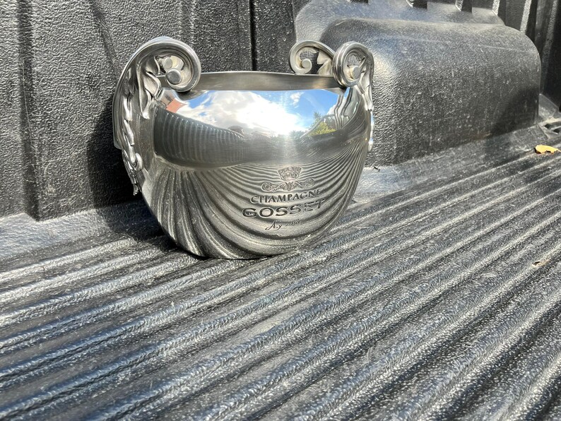 Puede incluir: Una cubitera de champ&aacute;n plateada con asas ornamentadas y un dise&ntilde;o con textura de concha. La cubitera tiene las palabras "CHAMPAGNE GOSSET" impresas en la parte delantera. La cubitera est&aacute; sobre una superficie texturizada.