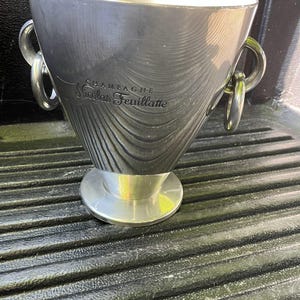Puede incluir: Una cubitera de champ&aacute;n plateada con las palabras "CHAMPAGNE Nicolas Feuillatte" grabadas en el lateral. La cubitera tiene dos asas de anillo y una base circular. La cubitera es de metal y tiene una superficie texturizada.