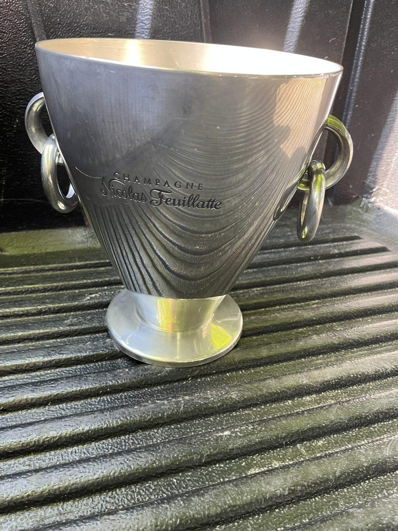 Puede incluir: Una cubitera de champ&aacute;n plateada con las palabras "CHAMPAGNE Nicolas Feuillatte" grabadas en el lateral. La cubitera tiene dos asas de anillo y una base circular. La cubitera es de metal y tiene una superficie texturizada.