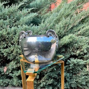 Puede incluir: Una cubeta de champ&aacute;n plateada con asas ornamentadas, que refleja la vegetaci&oacute;n circundante. La cubeta se encuentra sobre un soporte de metal dorado con una tapa de cristal. El texto "CHAMPAGNE GOSSET Ay 1584" es visible en la cubeta.