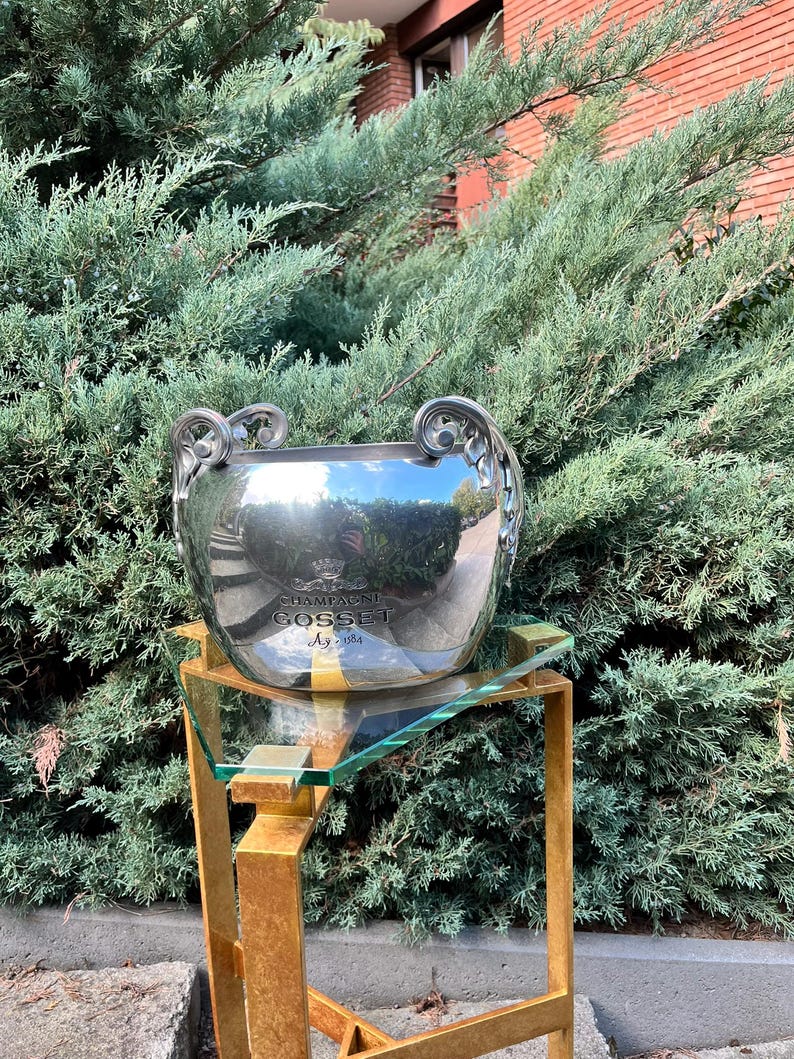 Puede incluir: Una cubeta de champ&aacute;n plateada con asas ornamentadas, que refleja la vegetaci&oacute;n circundante. La cubeta se encuentra sobre un soporte de metal dorado con una tapa de cristal. El texto "CHAMPAGNE GOSSET Ay 1584" es visible en la cubeta.