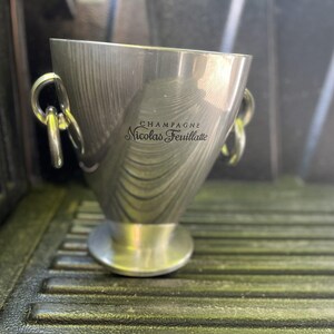Puede incluir: Una cubeta de champ&aacute;n de acero inoxidable con dos asas de anillo. La cubeta tiene una parte superior ancha y una base estrecha. El texto "CHAMPAGNE Nicolas Feuillatte" est&aacute; impreso en el lateral.