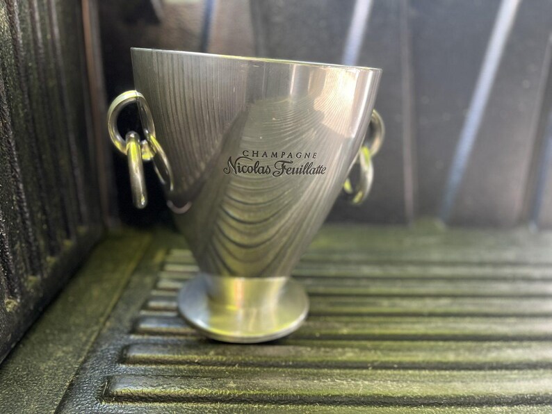 Puede incluir: Una cubeta de champ&aacute;n de acero inoxidable con dos asas de anillo. La cubeta tiene una parte superior ancha y una base estrecha. El texto "CHAMPAGNE Nicolas Feuillatte" est&aacute; impreso en el lateral.