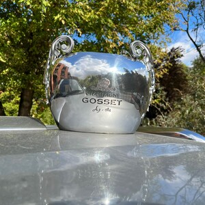 Puede incluir: Un cubo de champ&aacute;n plateado con la inscripci&oacute;n "CHAMPAGNE GOSSET" y "Ag. 1584". El cubo tiene dos asas ornamentadas y refleja el entorno. Se encuentra sobre una superficie plateada.