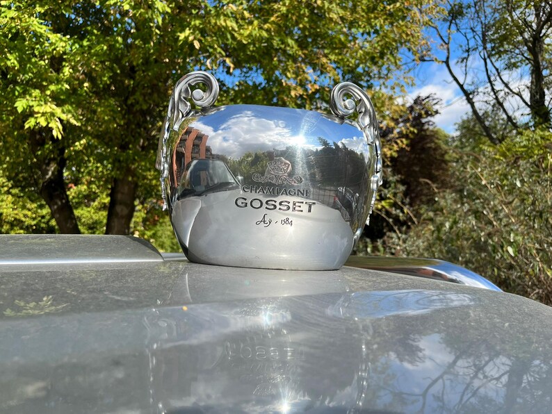 Puede incluir: Un cubo de champ&aacute;n plateado con la inscripci&oacute;n "CHAMPAGNE GOSSET" y "Ag. 1584". El cubo tiene dos asas ornamentadas y refleja el entorno. Se encuentra sobre una superficie plateada.