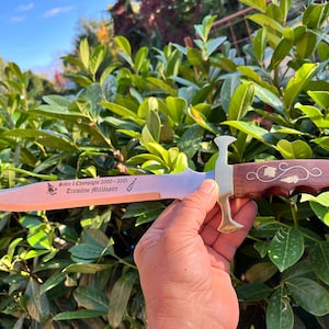 Könnte beinhalten: Ein dekoratives Messer mit polierter Stahlklinge und Holzgriff. Der Griff hat Messingakzente und ein verziertes Design. Die Klinge hat geätzten Text. Das Messer wird vor einem Hintergrund aus grünem Laub gehalten.