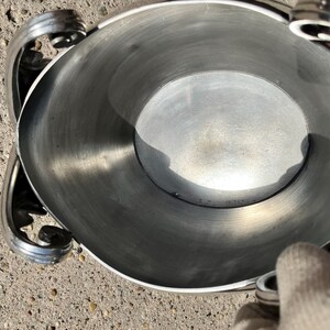 Puede incluir: Un cuenco de metal plateado con asas decorativas. El cuenco tiene una abertura circular ancha y un interior liso y reflectante. Las asas presentan un dise&ntilde;o de volutas. El cuenco probablemente se utiliza para servir o exhibir.