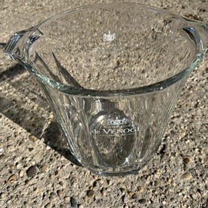 May include: A clear glass champagne bucket with a wide, fluted body and two handles. The bucket has the words "Champagne de Venoge" printed on the front. The bucket is designed to hold a bottle of champagne and ice.