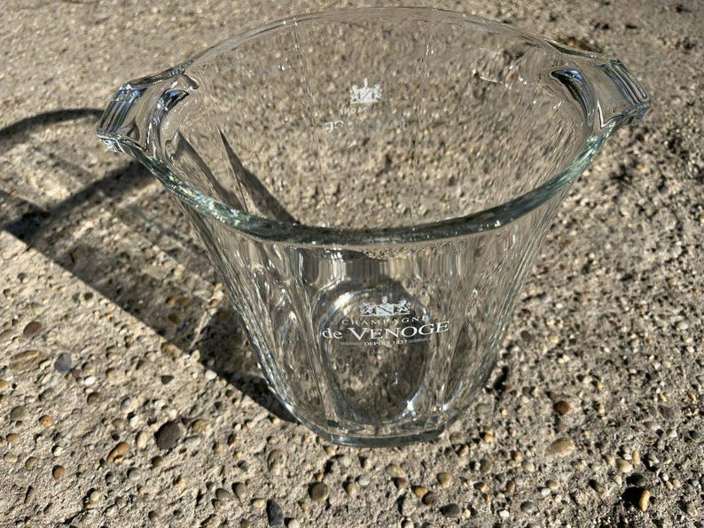 May include: A clear glass champagne bucket with a wide, fluted body and two handles. The bucket has the words "Champagne de Venoge" printed on the front. The bucket is designed to hold a bottle of champagne and ice.