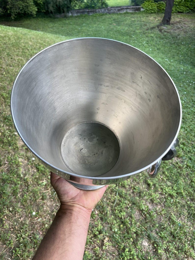 Puede incluir: Una cubitera grande de metal plateado con una abertura circular ancha y una base circular m&aacute;s peque&ntilde;a. La cubitera tiene un acabado pulido y dos asas curvas. El interior es liso y reflectante.