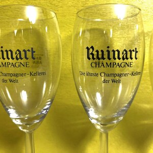 May include: Clear champagne flutes with the "Ruinart CHAMPAGNE" logo in black. The text "Die &auml;lteste Champagner-Kellerei der Welt" is also visible. The flutes are on a yellow background.