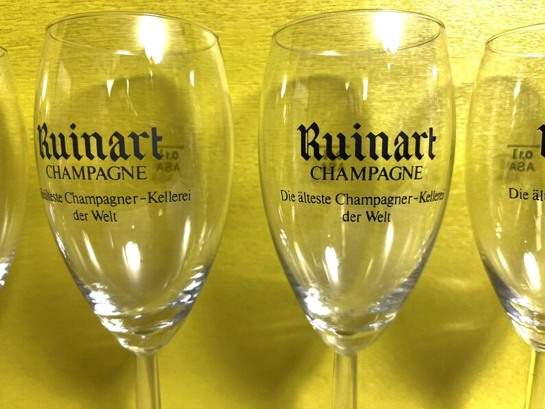 May include: Clear champagne flutes with the "Ruinart CHAMPAGNE" logo in black. The text "Die &auml;lteste Champagner-Kellerei der Welt" is also visible. The flutes are on a yellow background.