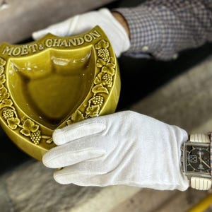 K&ouml;nnte beinhalten: Ein vintage, olivgr&uuml;nes Keramik-Aschenbecher in Schildform, gepr&auml;gt mit dem Logo "MO&Euml;T & CHANDON" und Weinreben-Details. Der Aschenbecher wird von behandschuhten H&auml;nden gehalten. Die Uhr am Handgelenk hat ein rechteckiges Zifferblatt.