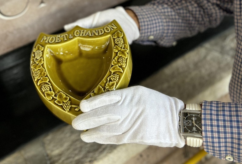 K&ouml;nnte beinhalten: Ein vintage, olivgr&uuml;nes Keramik-Aschenbecher in Schildform, gepr&auml;gt mit dem Logo "MO&Euml;T & CHANDON" und Weinreben-Details. Der Aschenbecher wird von behandschuhten H&auml;nden gehalten. Die Uhr am Handgelenk hat ein rechteckiges Zifferblatt.