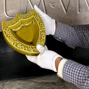 K&ouml;nnte beinhalten: Ein schildf&ouml;rmiger, olivgr&uuml;ner Keramik-Aschenbecher mit dem Logo "MO&Euml;T & CHANDON". Der Aschenbecher hat einen dekorativen Rand aus Weinreben und wird von behandschuhten H&auml;nden gehalten. Der Hintergrund zeigt gemei&szlig;elten Stein.