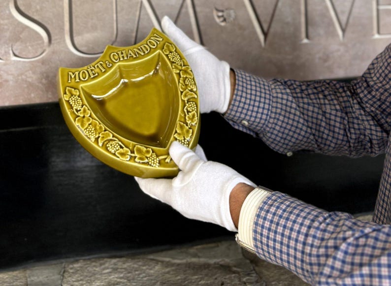 K&ouml;nnte beinhalten: Ein schildf&ouml;rmiger, olivgr&uuml;ner Keramik-Aschenbecher mit dem Logo "MO&Euml;T & CHANDON". Der Aschenbecher hat einen dekorativen Rand aus Weinreben und wird von behandschuhten H&auml;nden gehalten. Der Hintergrund zeigt gemei&szlig;elten Stein.