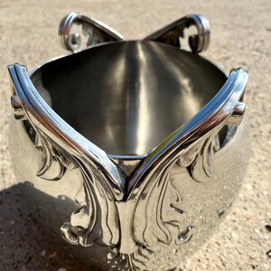 Puede incluir: Cubeta de hielo de metal plateado con asas ornamentadas en forma de hojas y adornos decorativos. La cubeta tiene un cuerpo redondeado y un interior pulido. El dise&ntilde;o sugiere un estilo vintage o antiguo.