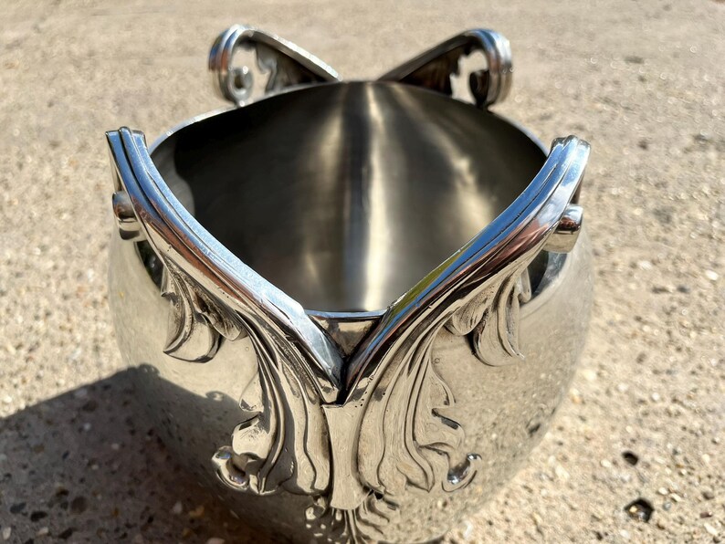 Puede incluir: Cubeta de hielo de metal plateado con asas ornamentadas en forma de hojas y adornos decorativos. La cubeta tiene un cuerpo redondeado y un interior pulido. El dise&ntilde;o sugiere un estilo vintage o antiguo.