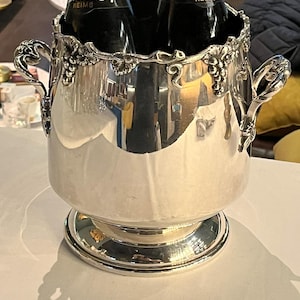 Peut inclure: Seau à champagne argenté avec deux bouteilles de champagne à l'intérieur. Le seau est orné de poignées et d'un motif de vigne sur le bord. Les bouteilles de champagne ont des étiquettes dorées.
