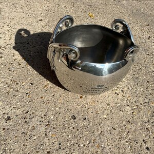 Puede incluir: Un cubo de champ&aacute;n de metal plateado brillante con asas ornamentadas. El cubo tiene una forma redondeada y una superficie pulida, que refleja el entorno. Las asas presentan un dise&ntilde;o de volutas decorativas. El cubo se encuentra sobre una superficie de hormig&oacute;n texturizado.