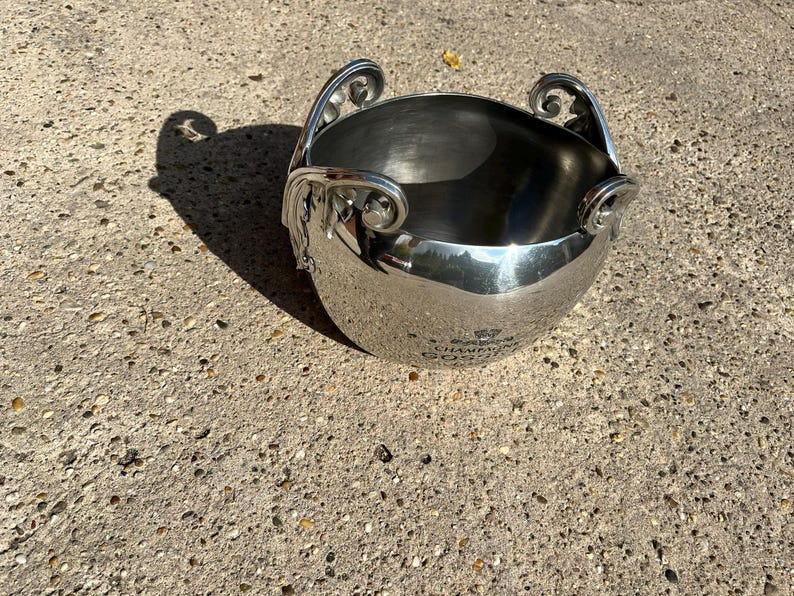 Puede incluir: Un cubo de champ&aacute;n de metal plateado brillante con asas ornamentadas. El cubo tiene una forma redondeada y una superficie pulida, que refleja el entorno. Las asas presentan un dise&ntilde;o de volutas decorativas. El cubo se encuentra sobre una superficie de hormig&oacute;n texturizado.