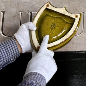 K&ouml;nnte beinhalten: Ein schildf&ouml;rmiger, olivgr&uuml;ner Keramik-Teller mit cremefarbenem Rand wird von behandschuhten H&auml;nden gehalten. Der Teller hat eine gl&auml;nzende Oberfl&auml;che und ein strukturiertes Inneres. Der Hintergrund zeigt in Stein gemei&szlig;elte Buchstaben.