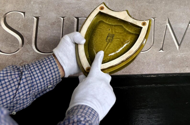 K&ouml;nnte beinhalten: Ein schildf&ouml;rmiger, olivgr&uuml;ner Keramik-Teller mit cremefarbenem Rand wird von behandschuhten H&auml;nden gehalten. Der Teller hat eine gl&auml;nzende Oberfl&auml;che und ein strukturiertes Inneres. Der Hintergrund zeigt in Stein gemei&szlig;elte Buchstaben.